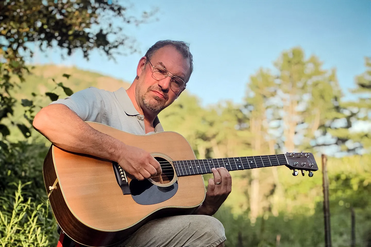 Jesse Sterling Harrison seated outdoors in a sunlit backyard, playing an acoustic guitar, wearing a cap and glasses.